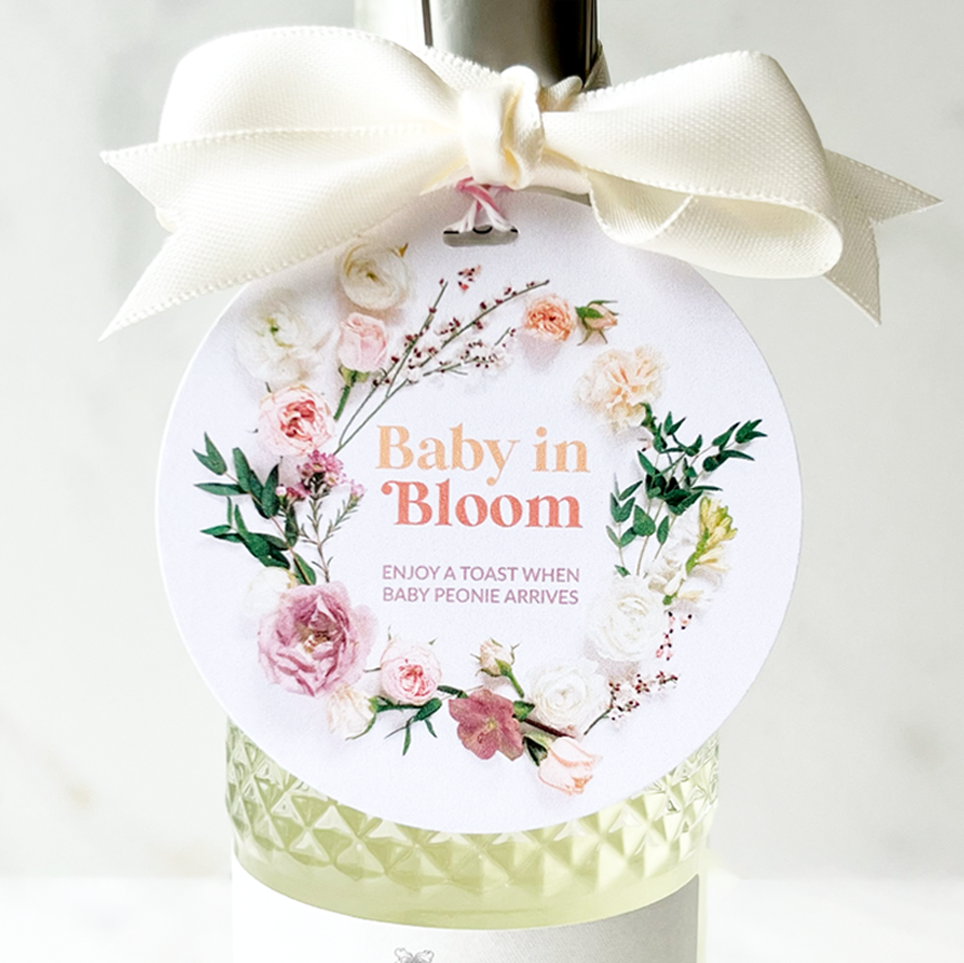 round decorative floral tag with flowers on white card stock reads baby in bloom enjoy a toast when baby peonie arrives tied on mini bottle of champagne