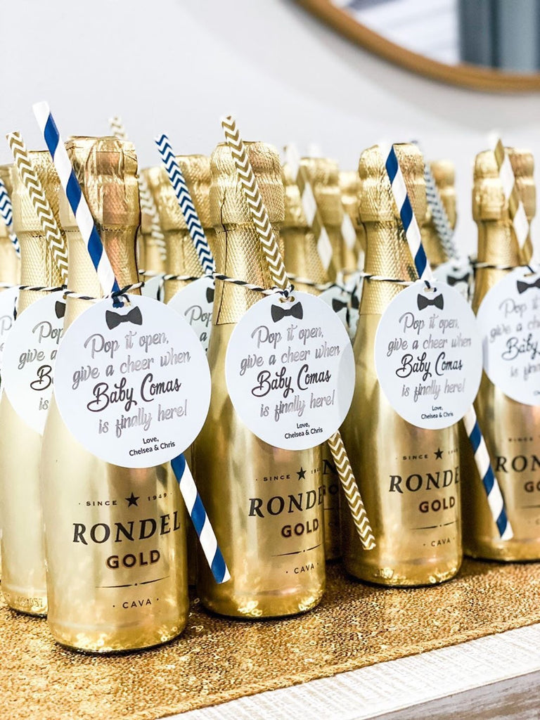 bright gold bottles of mini champagne in decorative rows on display for baby shower guests. small tags are tied around the bottles which read pop it open send a cheer when baby comas is finally here. A paper straw is also included